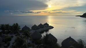 A beach with thatched-roof huts, palm trees, and a calm sea at sunset. - 2-bedroom apartment in Blue Bay Resort (Willemstad)