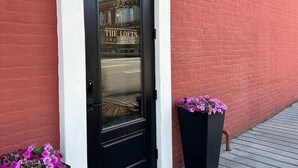 Una puerta negra con la señalización 'The Lofts', un muro de ladrillo rojo y dos macetas negras con flores rosadas.