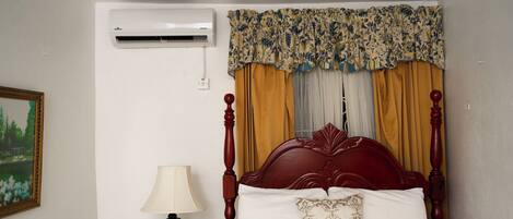 A bedroom with a wooden bed, a nightstand with a lamp, a wall-mounted air conditioner, and a framed picture on the wall.