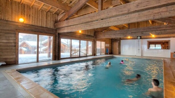 An indoor swimming pool with wooden beams, large windows, and a view of snow-covered buildings outside. - Eden Criou - Chalet sleeps 7 to 9 - pool/sauna (Morillon)