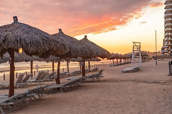A beach with thatched umbrellas and sun loungers, a lifeguard tower, and a high-rise building in the background.