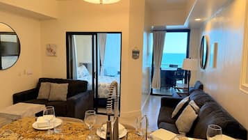 Una habitación de hotel moderna con zona de comedor, sofá, amplio ventanal con vista al mar y ventilador de techo.