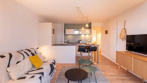 A modern living room with a sofa, a coffee table, and a TV. There is a kitchen area in the background with a dining table and chairs. - Carnac - 3-room apartment - 50m² - Wifi - Pool (CARNAC)