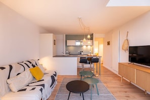 A modern living room with a sofa, a coffee table, and a TV. There is a kitchen area in the background with a dining table and chairs. - Carnac - 3-room apartment - 50m² - Wifi - Pool (CARNAC)