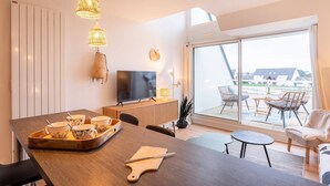 A modern living room with a wooden dining table, a TV, and a balcony with wicker chairs. - Carnac - 3-room apartment - 50m² - Wifi - Pool (CARNAC)