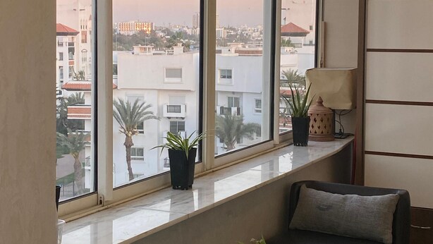Un salon moderne doté d’une grande fenêtre donnant sur un paysage urbain. La pièce est aménagée avec un canapé en cuir noir, une petite table basse et deux plantes en pot placées sur le rebord de la fenêtre.