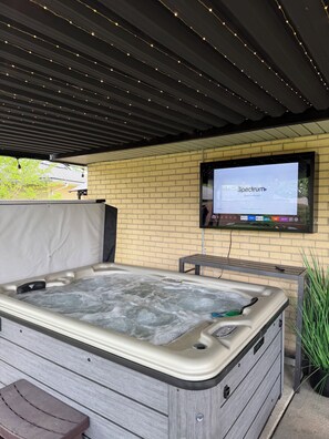 A spa area with a hot tub, a television mounted on a brick wall, and a ceiling with string lights. - Fire Pit, Private Hot Tub + Workspace - Sleeps 7 (Dayton)