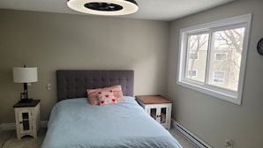 A bedroom with a bed, a nightstand, a lamp, a heater, and a window with a view of buildings.