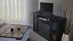 A living room with a wooden coffee table, a dark entertainment unit with a flat-screen TV, a vase with branches, and a window with sheer curtains.