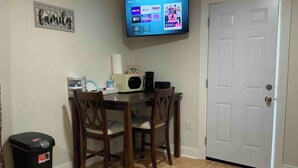 Ein Essbereich mit Tisch und Stühlen, einem an der Wand befestigten Fernseher, einem Mülleimer und einer Tür.