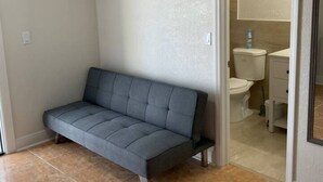 Ein Zimmer mit einer grauen Couch, einer weißen Toilette und einem durchgehenden Türspalt sichtbaren Badezimmer.