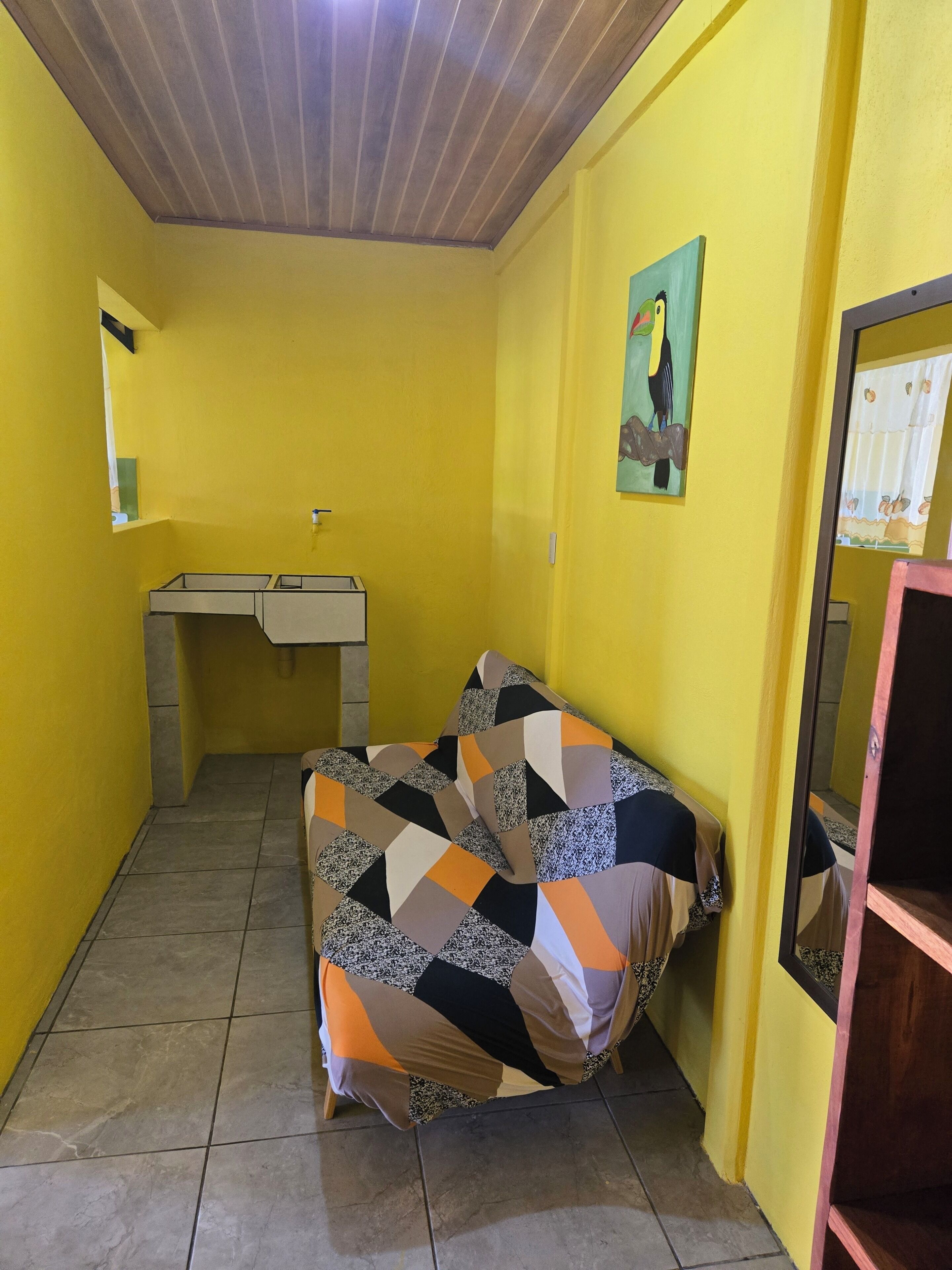 A small room with yellow walls, a wooden ceiling, a bed with a patterned blanket, a small desk with a mirror, and a wall-mounted painting.