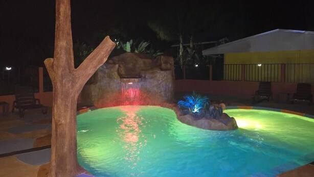 An outdoor swimming pool with a waterfall feature and illuminated plants. - Rusticas Dani & Fam (La Fortuna)