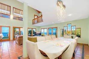 A spacious dining area with a high ceiling, large windows offering ocean views, and a balcony with wooden railings. - Kona Oceanview Villa - Luxury Group House with Pool, Hot Tub, and Coastline View (Captain Cook)
