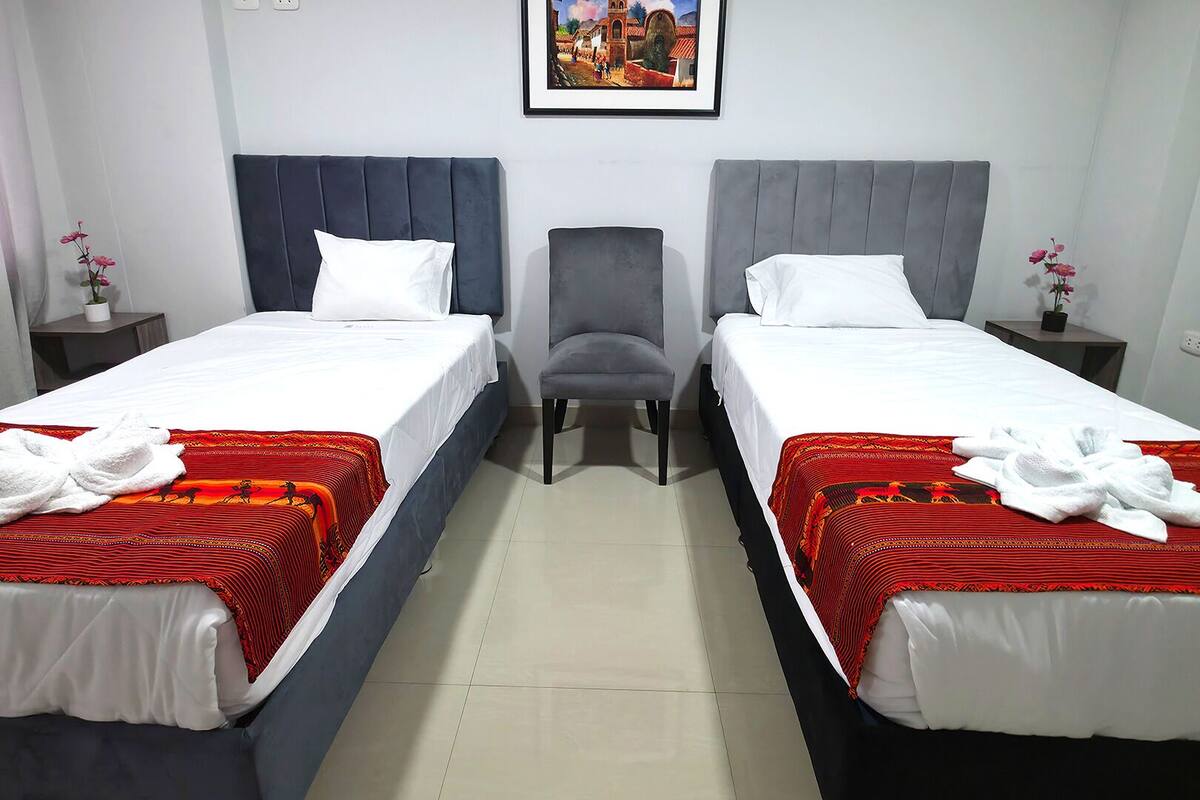 Camera d'albergo con due letti, ognuno con coperta a motivi rossi e neri e un asciugamano bianco. Al centro, una poltrona grigia.