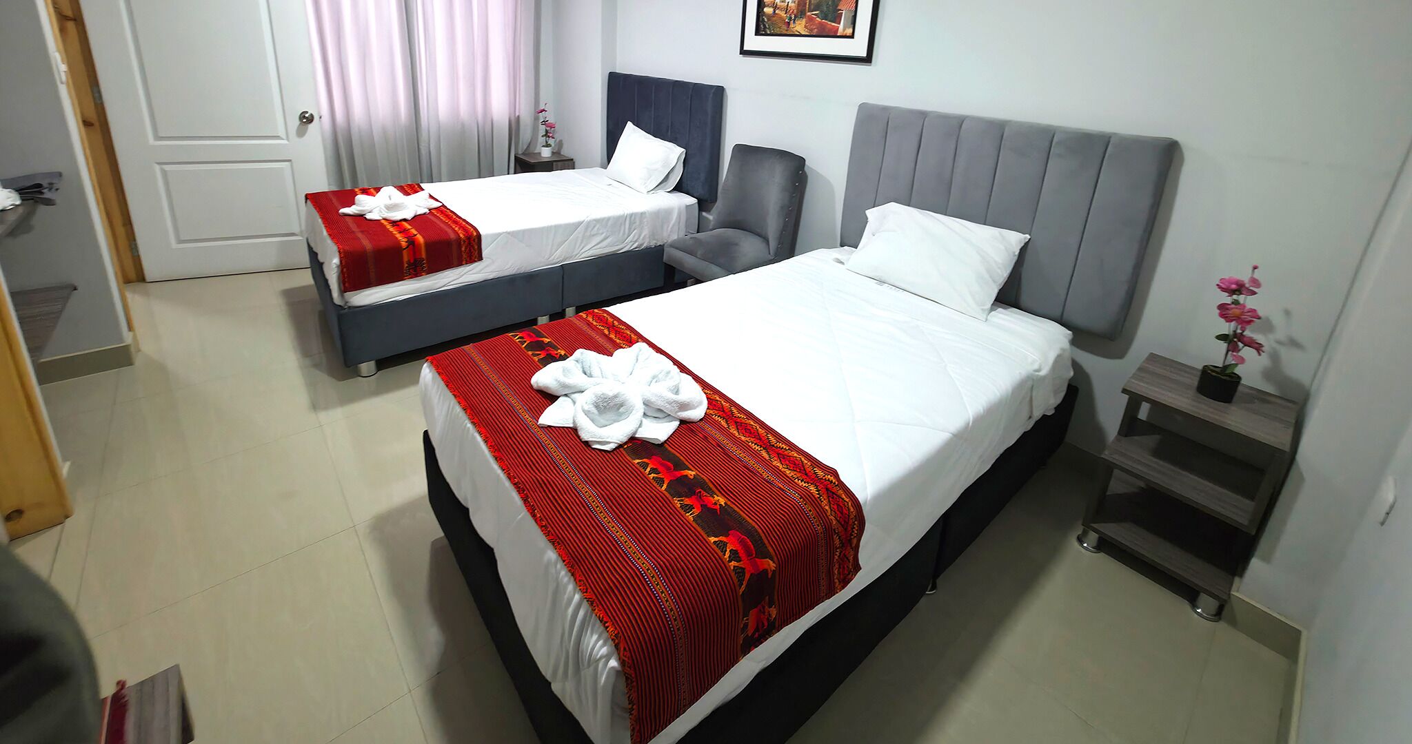 A hotel room with two beds, each with a red and white bedspread and white pillows. There is a grey headboard on the larger bed and a chair next to it. A small wooden nightstand with a potted plant is on the right side of the room.