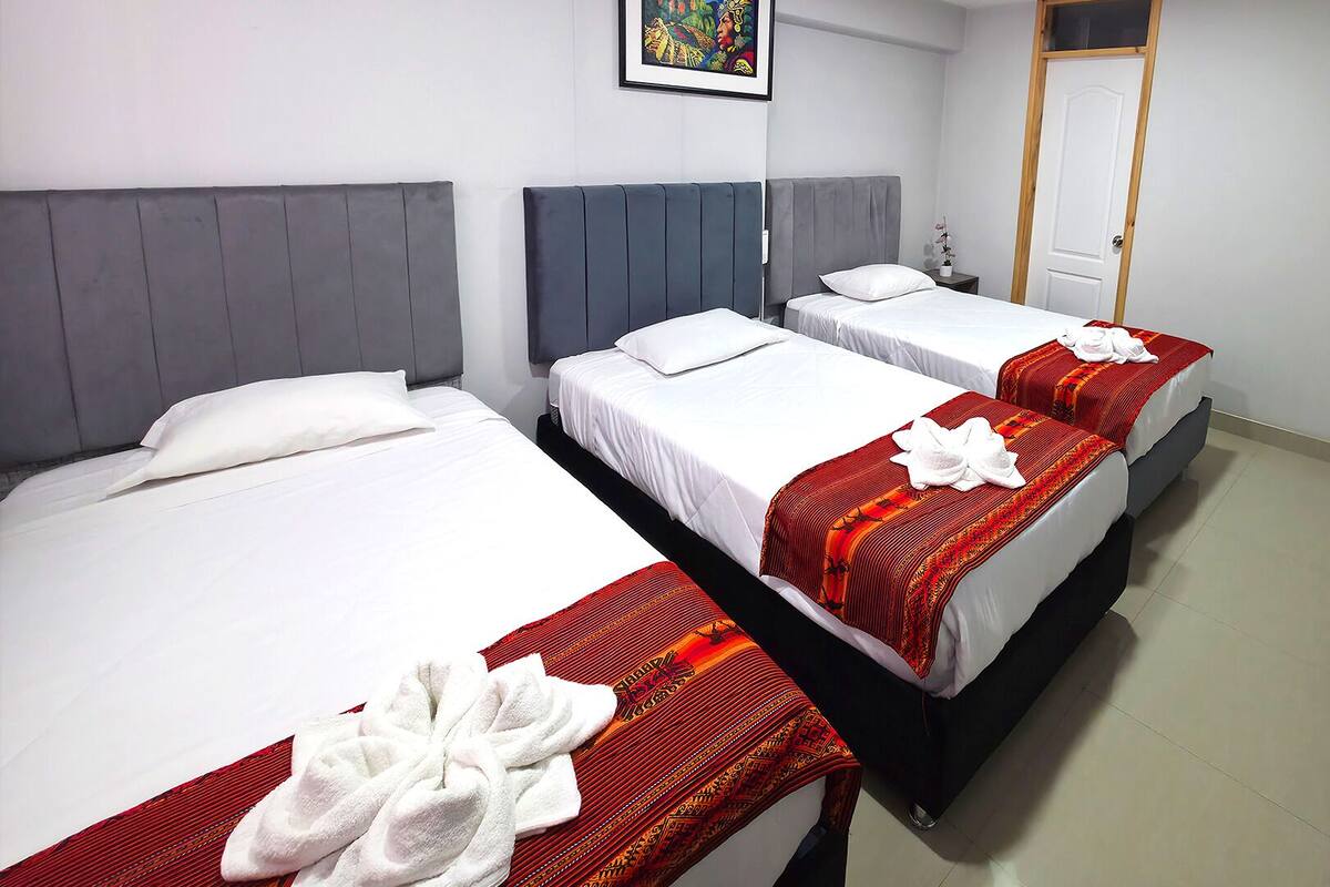Una camera d'albergo con tre letti singoli, ognuno con una coperta bianca e una coperta a motivi rossi. Si vede un quadro appeso al muro, una porta in legno e un tavolino con un vaso di fiori.