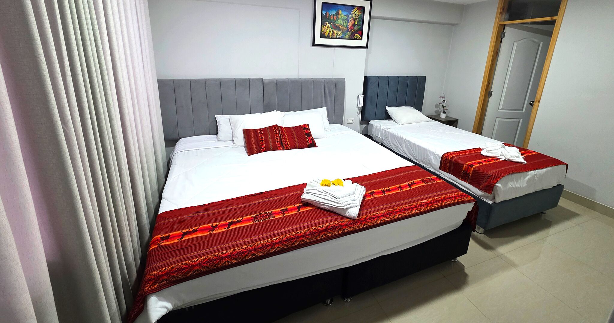 A hotel room with two beds, each with a grey headboard, white bedding, and red decorative pillows. A red patterned blanket is draped over the larger bed, and a white towel is folded on it.