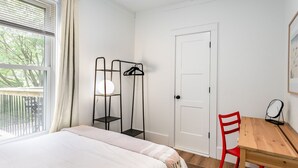 Een slaapkamer met een bed, een houten tafel, een rode stoel, een deur en een raam met gordijnen.