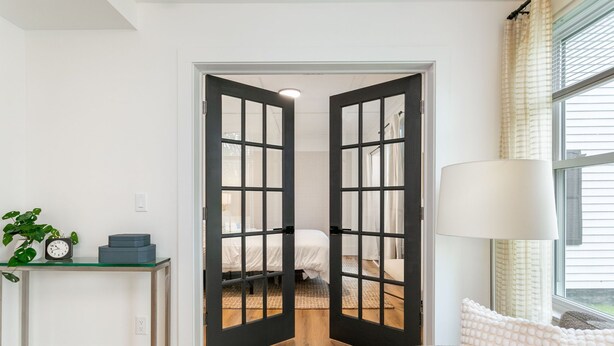Een kamer met een bed, een tafel met een klok, een lamp en een raam met gordijnen.