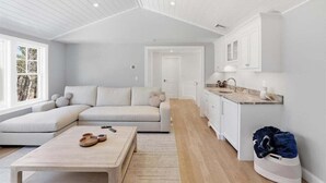 A modern interior with a sofa, a coffee table, a kitchen area, and a view through a window. - Olivia's Oasis: 006-HL - Stunning New Construction with Heated Pool & Hot Tub in Harwich (Harwich)