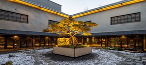 A courtyard with a central tree, stone pathways, and a building with large windows and warm lighting. - AT STAY (Cheonan)