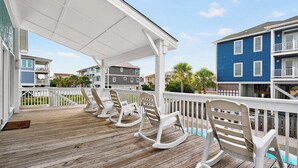 A wooden deck with white rocking chairs, a covered area, and a view of neighboring buildings. - Sea + Second | Pool & Short Walk to the Beach (Ocean Isle Beach)