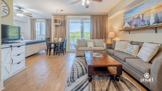A spacious living room with a sofa, coffee table, and a television. There is a balcony with curtains and a painting of a beach scene. - “Sun-kissed” condo , Fantastic location in Baytown Wharf,  Sandestin. (Miramar Beach)