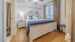 A bedroom with a wooden bed, a nightstand, a ceiling fan, and a window with curtains. - “Sun-kissed” condo , Fantastic location in Baytown Wharf,  Sandestin. (Miramar Beach)