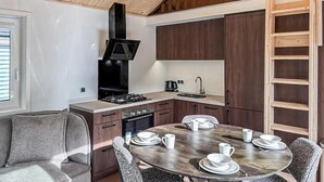 Een kleine keuken met een fornuis, spoelbak en een zwarte afzuigkap. Er staat een eettafel met borden en kopjes, en een bank op de voorgrond.