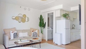 A modern living room with a sofa, coffee table, and a dining area. There is a kitchen with white cabinets and a refrigerator. A large plant is visible in the living room. - Oceanview Remodeled Studio in the heart of Waikiki, walk to shops, food, & beach (Honolulu)