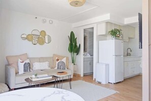Ein modernes Wohnzimmer mit Sofa, Couchtisch und Essbereich. Es gibt eine Küche mit weißen Schränken und einem Kühlschrank. Eine große Pflanze ist im Wohnzimmer zu sehen.
