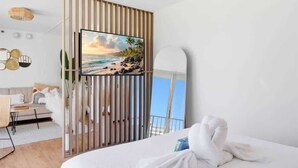 A hotel room with a bed, a view of the beach, a wooden partition, and a wall-mounted TV. - Oceanview Remodeled Studio in the heart of Waikiki, walk to shops, food, & beach (Honolulu)