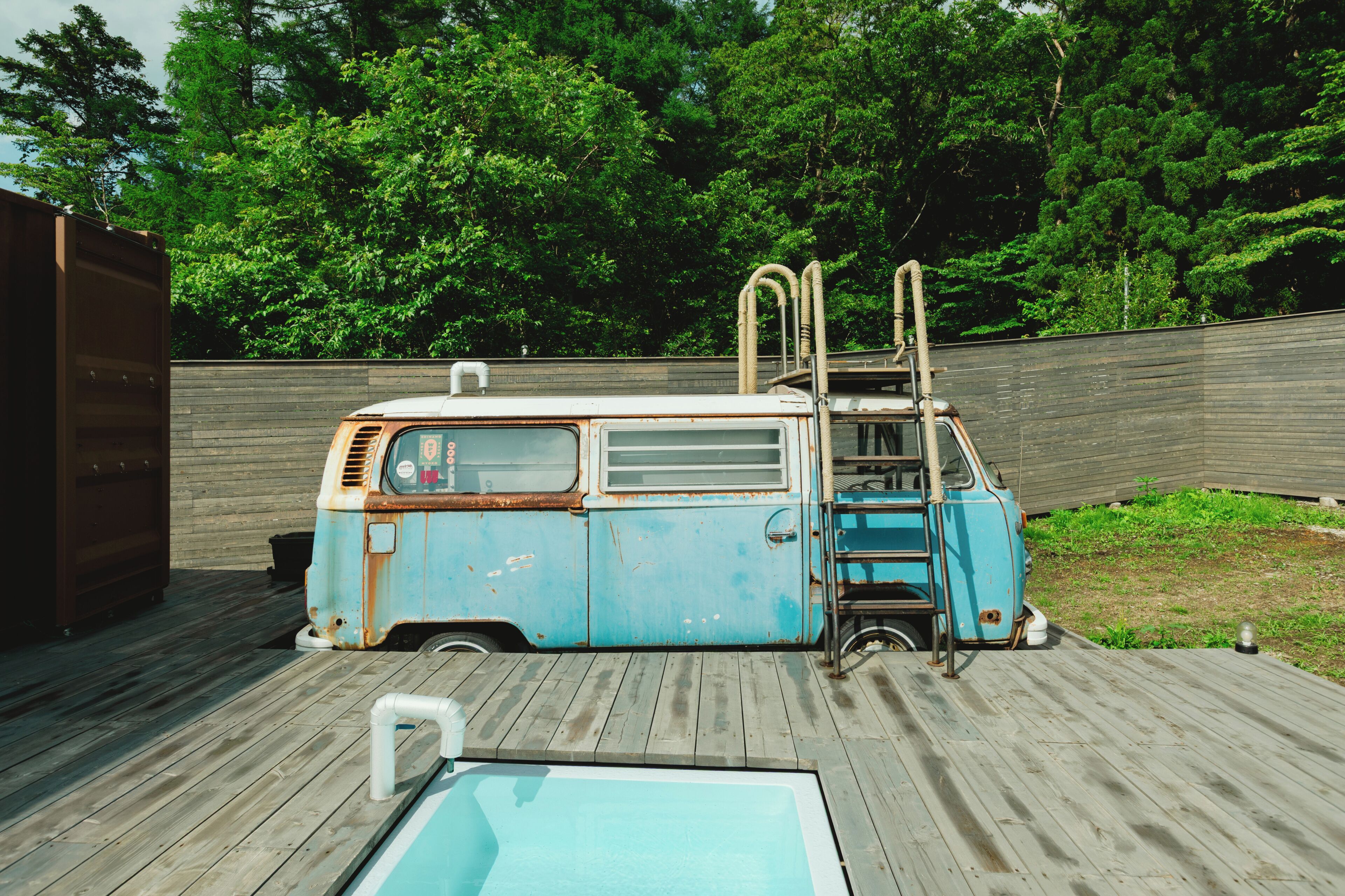 A blue van with a ladder, a wooden deck, and a swimming pool.