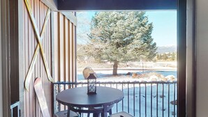 Ein Balkon mit rundem Tisch und Laterne, mit Blick auf eine verschneite Landschaft mit Bäumen und einem fernen Gebirge.