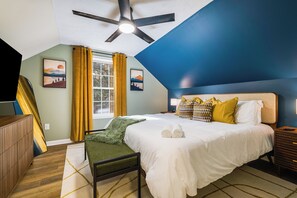 Un dormitorio con cama, ventilador de techo, ventana con cortinas amarillas y obras de arte en las paredes.