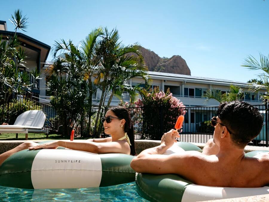 A pool area with inflatable loungers labeled "SUNNYLIFE," palm trees, and a building in the background.