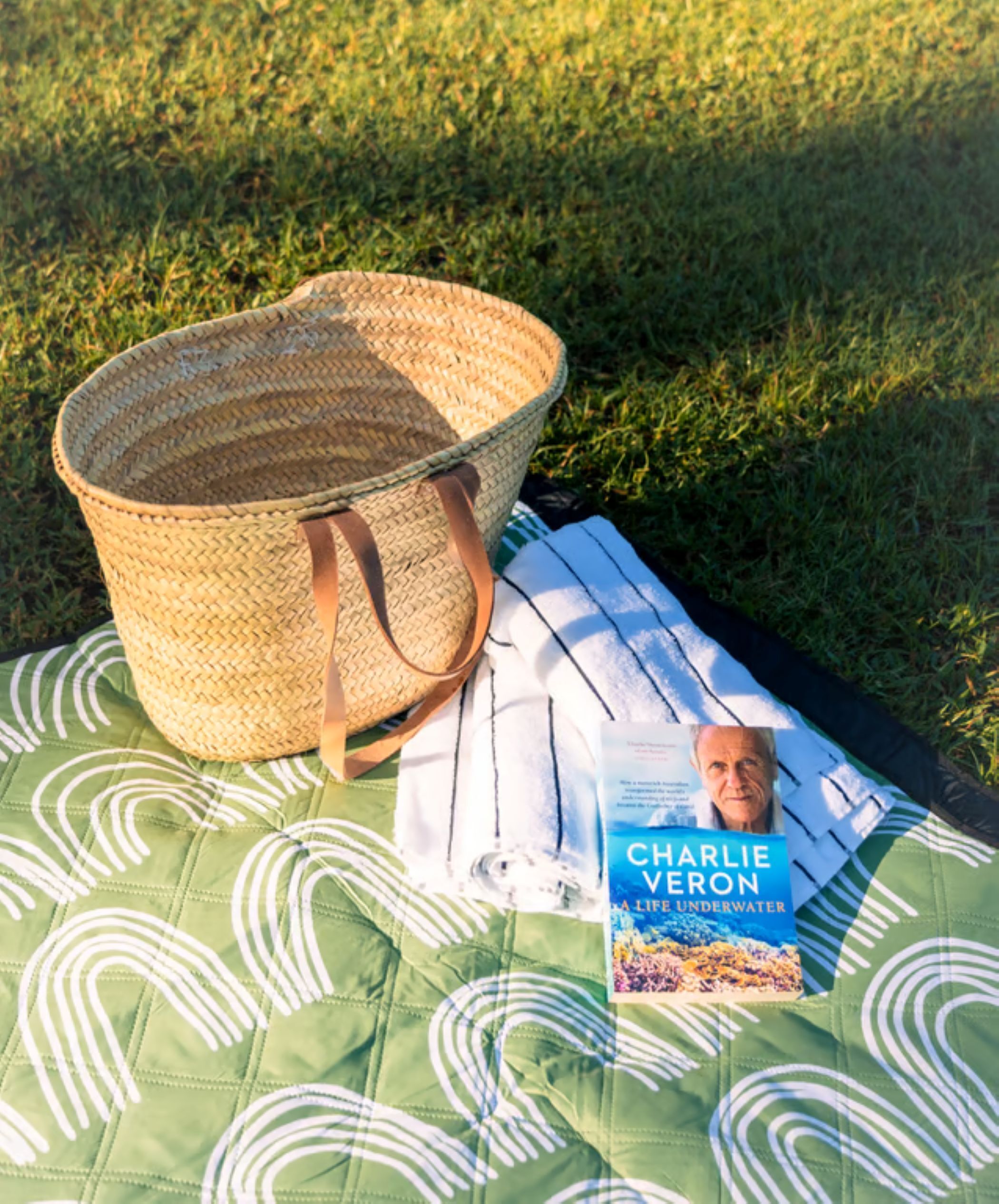 A picnic setup with a woven basket, a book titled 'Charlie Veron: A Life Underwater', and a green quilt with white patterns on a grassy field.