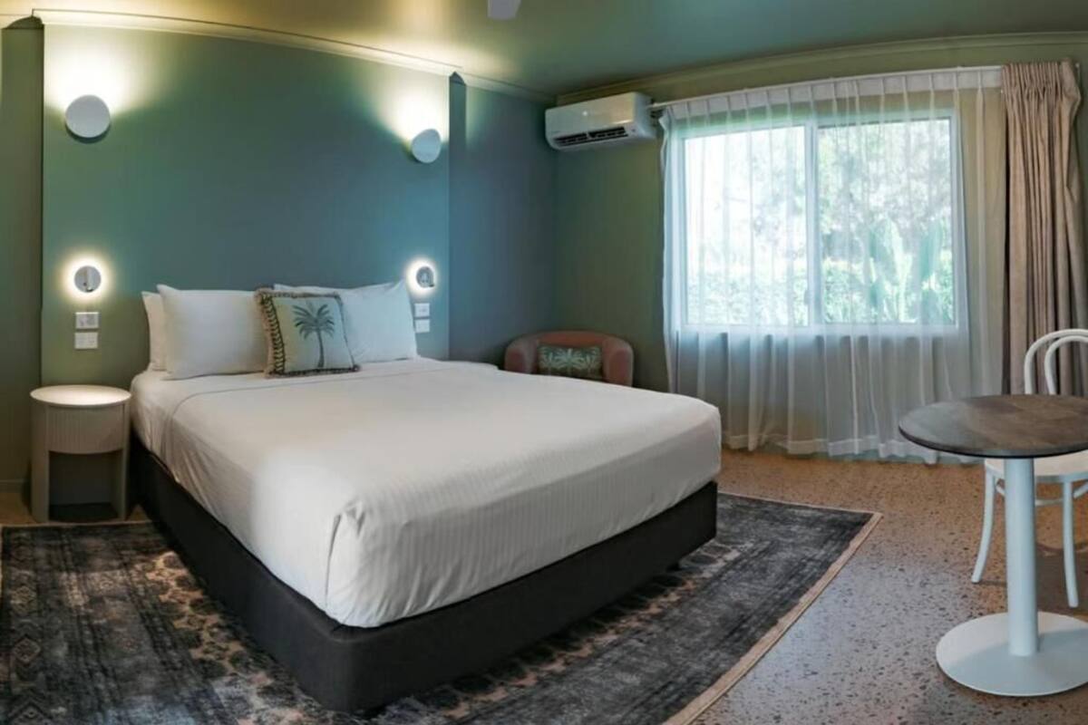 A bedroom with a bed, bedside lamps, an air conditioning unit, a window with curtains, and a round table.