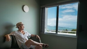 A man sitting on a couch in a room with a large window, a round wall clock, and a bed with a white bedspread. - Victoria Street Boutique Hotel (Townsville City)
