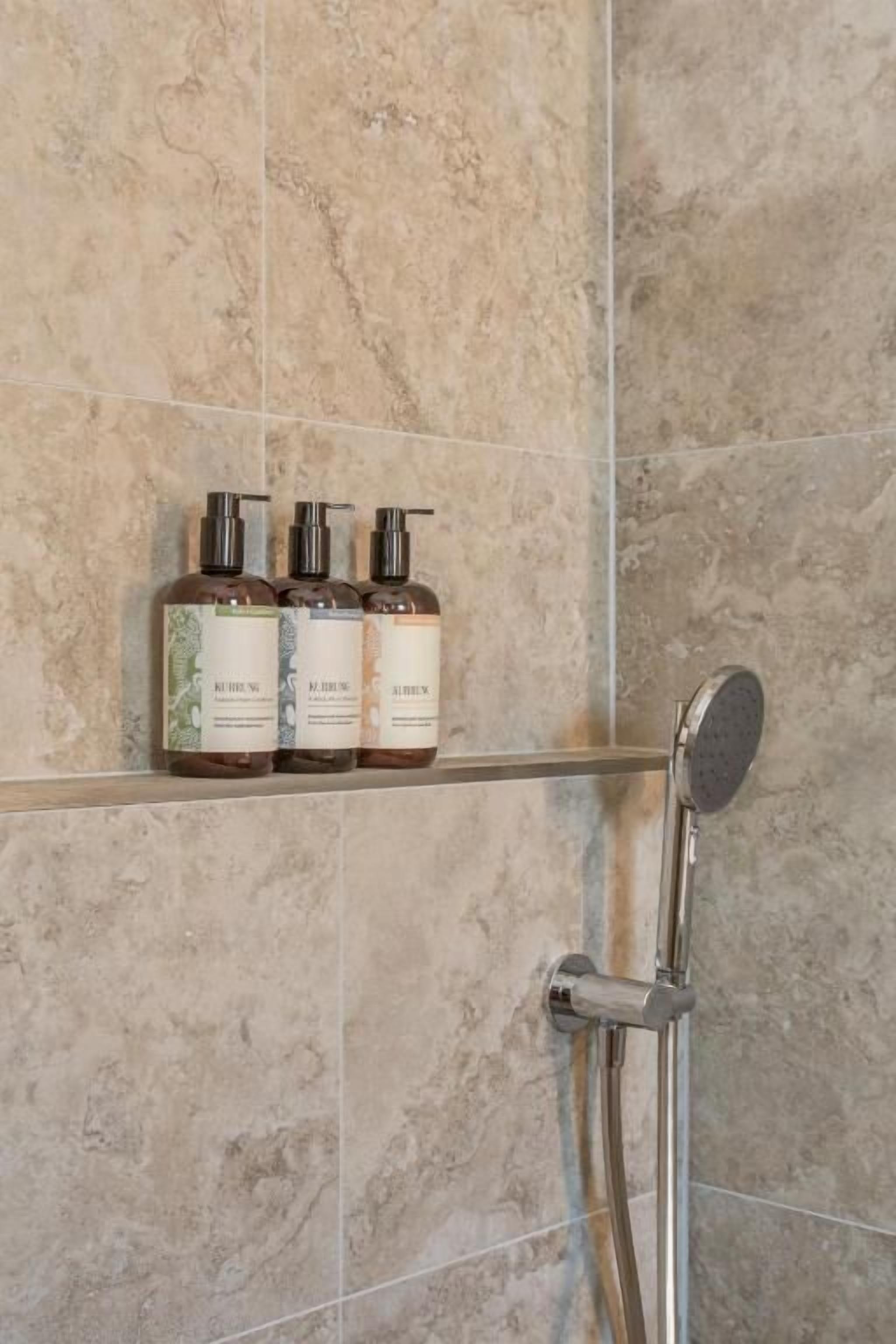 A bathroom with a shower, a shelf with three bottles of shampoo, and a tiled wall.