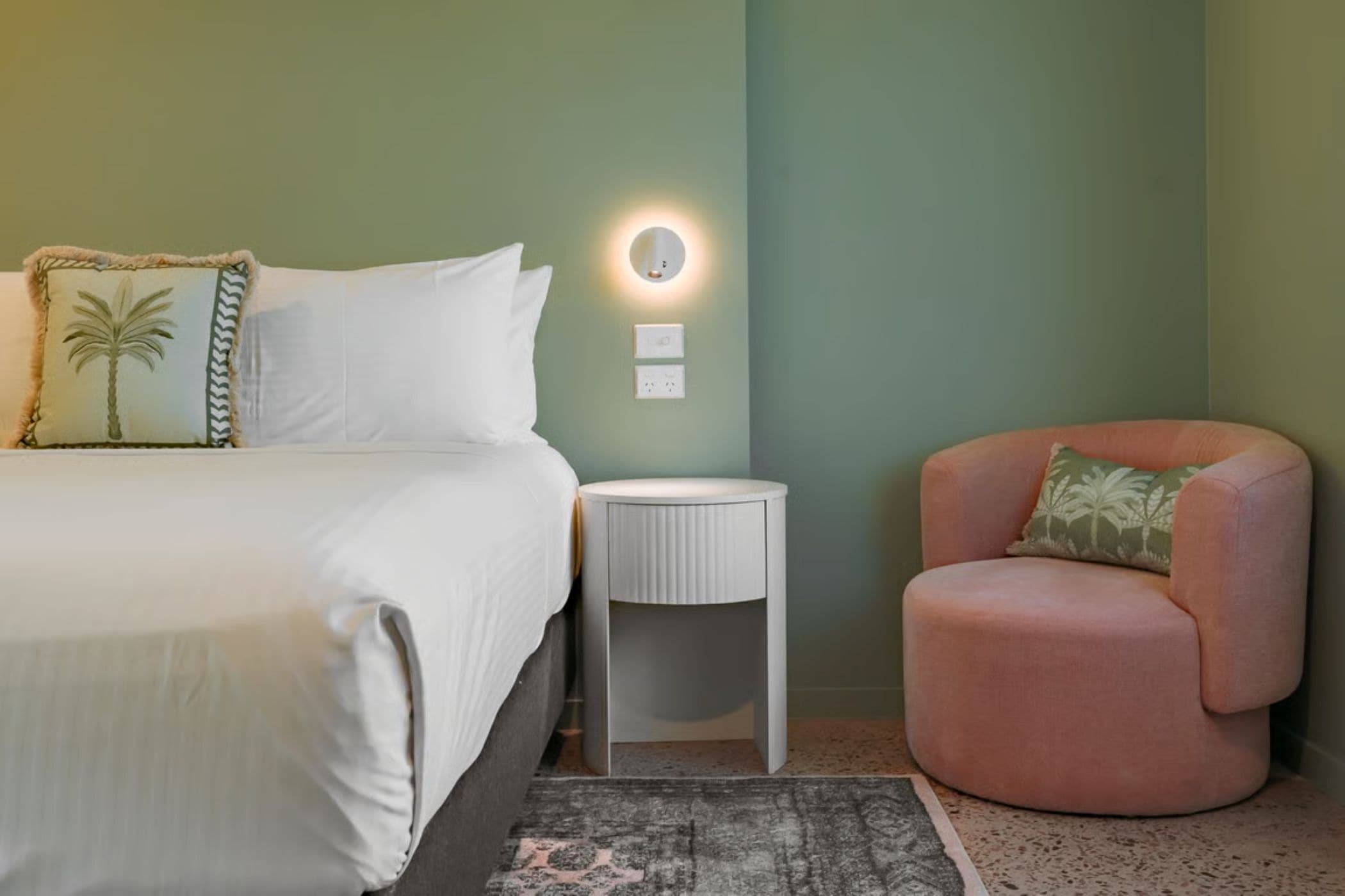 Un dormitorio con cama, una mesita de noche redonda, una silla y una lámpara fijada a la pared.