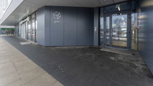Entrada de um edifício moderno com porta de vidro, um logotipo na parede e um caminho pavimentado.