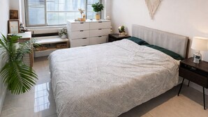 A bedroom with a large bed, a dresser, a nightstand, a wall-mounted dreamcatcher, and a window with blinds.