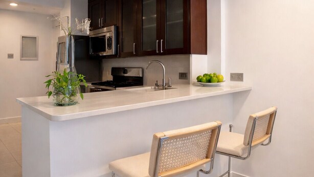 A modern kitchen with a white countertop, stainless steel appliances, dark wooden cabinets, and two bar stools with woven seats.