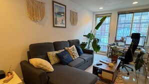 A modern living room with a grey sectional sofa, a small side table, a coffee table, and a desk with a laptop. There are two large windows providing natural light and a view of the outside.