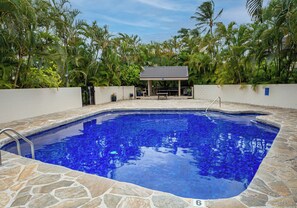 A swimming pool with a tiled deck, surrounded by a white wall and lush tropical vegetation. - Luxury Ocean-View 2BR Condo | Walk to Poipu Beach | Sleeps 6 (Koloa)