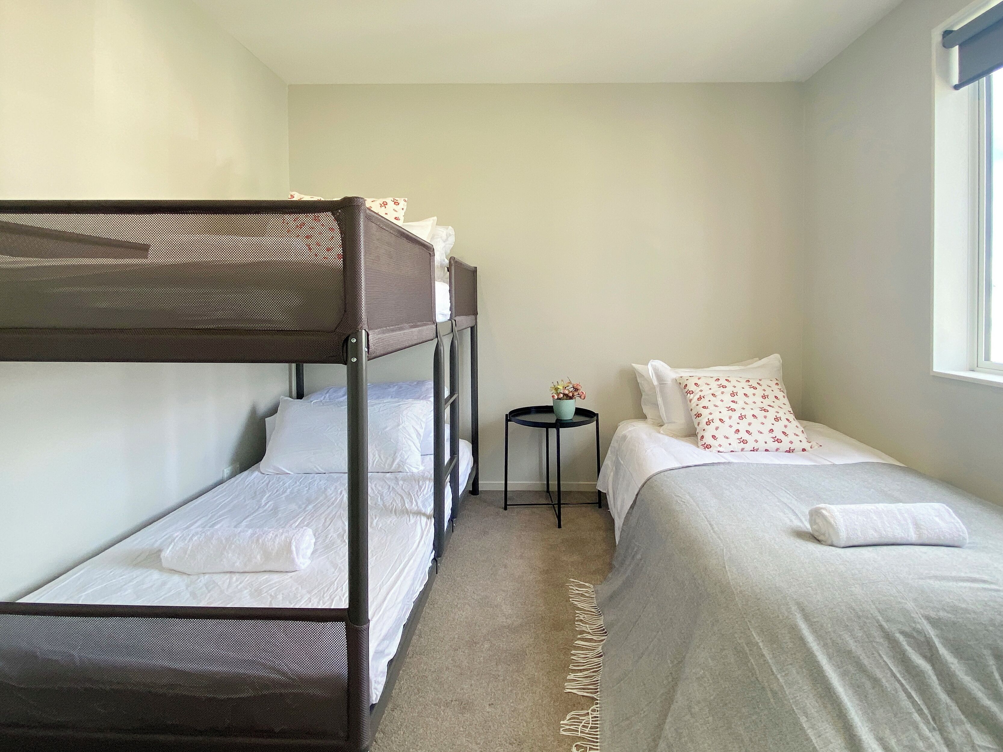 A compact room with bunk beds and a single bed, a small round table with a plant, and a window providing natural light.
