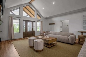 Un salon spacieux avec un plafond haut, de grandes fenêtres, un canapé, des poufs et une table basse.