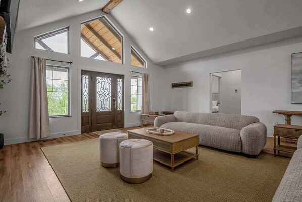 A spacious living room with a high ceiling, large windows, a sofa, ottomans, and a coffee table. - Pool | Sleeps 18 | Hot Tub |Firepit | Playset | (Williamstown)